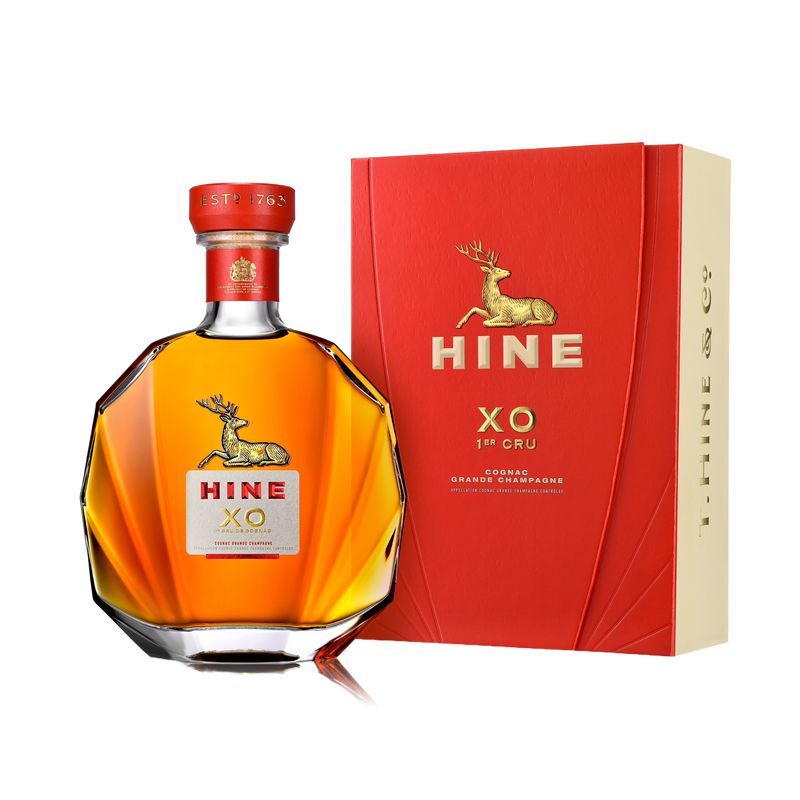A bottle of HINE XO 1er Cru Cognac by HINE Cognac with a gold deer logo and red label stands beside its matching red and gold box, both featuring the same design, all set against a white background.