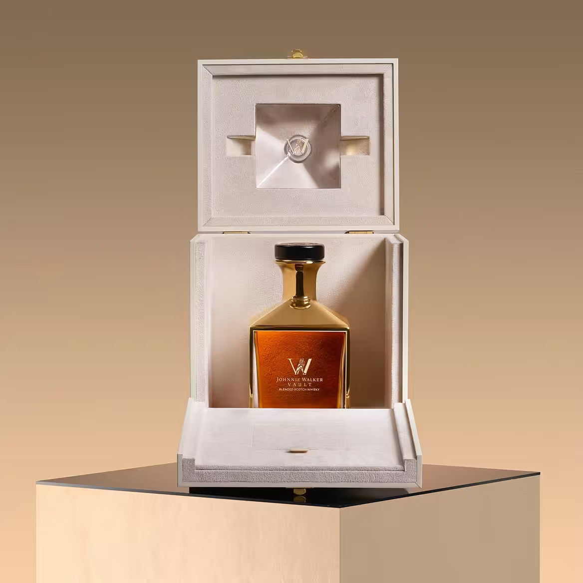 A luxurious bottle of Johnnie Walker Vault: The Couture Blend Olivier Rousteing 700ml is displayed in an elegant cream-colored box with a satin interior, set atop a reflective black and gold pedestal against a gradient beige background.