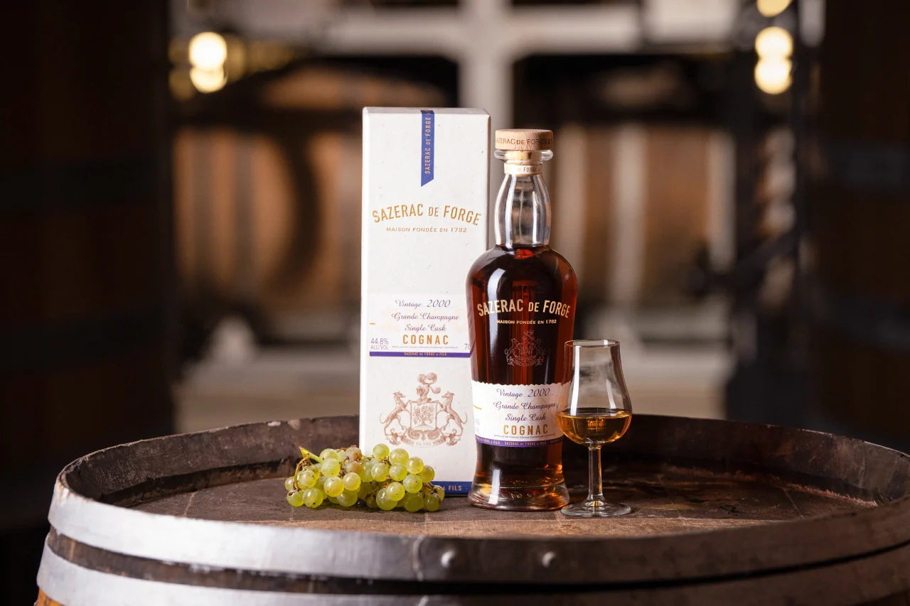 A bottle of Sazerac de Forge Grande Champagne Cognac Vintage 2000 by Sazerac, a glass of cognac, fresh green grapes, and the box are elegantly arranged on a wooden barrel with blurred barrels in the background.