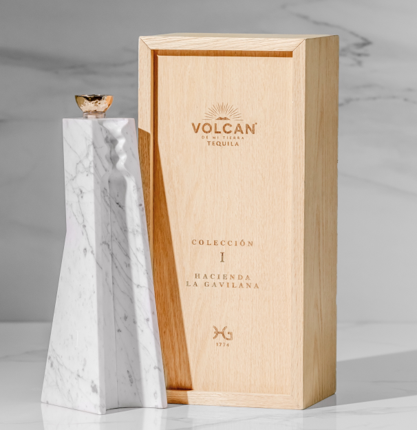 A white marble-patterned Volcan De Mi Tierra Coleccion I Hacienda La Gavilana Anejo Tequila bottle with a gold cap stands beside a light wooden box on a marble surface, both labeled with the Volcan De Mi Tierra brand.