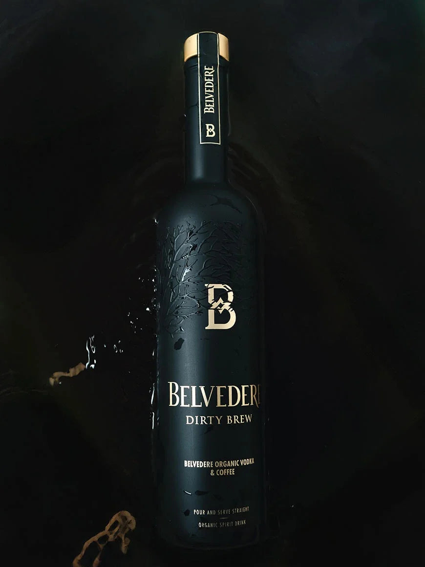 A matte black bottle of Belvedere Dirty Brew Organic Coffee Flavored Vodka by Belvedere Vodka, accented with gold lettering, rests on a dark surface amid coffee splashes. The label highlights “Organic Vodka & Coffee.”.