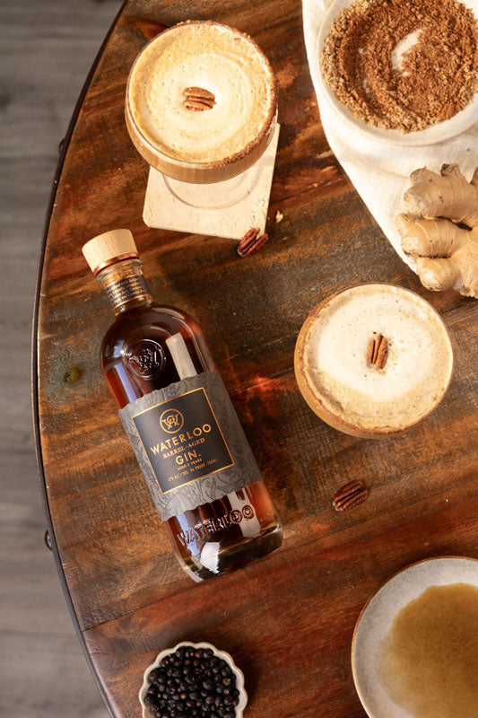 A bottle of Waterloo Barrel-Aged Gin by Waterloo Gin rests on a wooden table, surrounded by two creamy pecan cocktails, a bowl of brown sugar, ginger, and a dish of juniper berries.