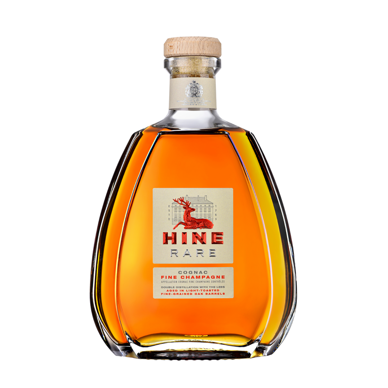 A clear, curvy bottle of HINE Cognac Rare by HINE Cognac features a wooden cork and a label with a red deer and gold accents. The amber liquid shows through the glass, all set against a plain white background.