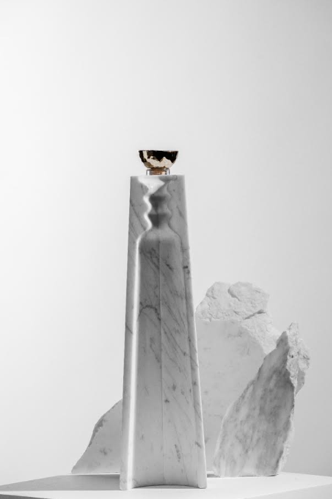 A small gold-rimmed cup sits atop a tall white marble pedestal, echoing the elegance of Volcan De Mi Tierra Coleccion I Hacienda La Gavilana Anejo Tequila, set before abstract white stone and a neutral wall.