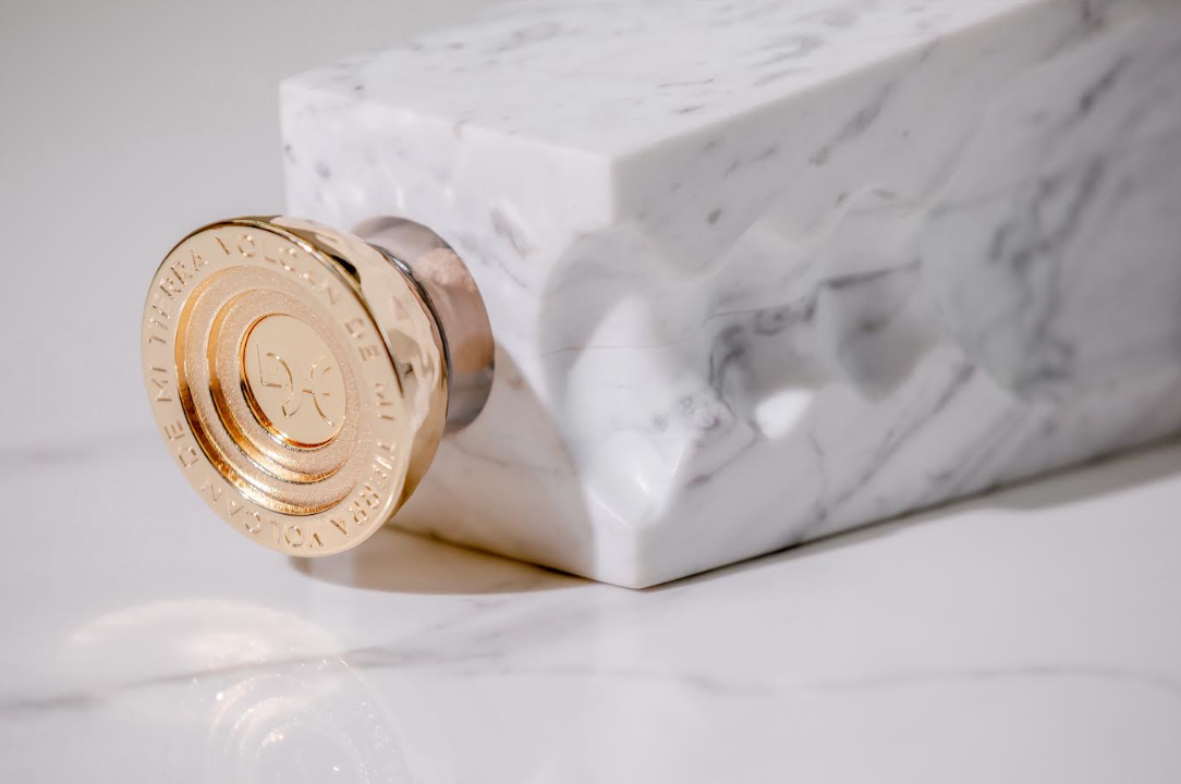 A gold-colored, round perfume bottle cap engraved with text sits atop a gray-veined white marble block, reflecting the elegance and refinement of Volcan De Mi Tierra Coleccion I Hacienda La Gavilana Anejo Tequila.