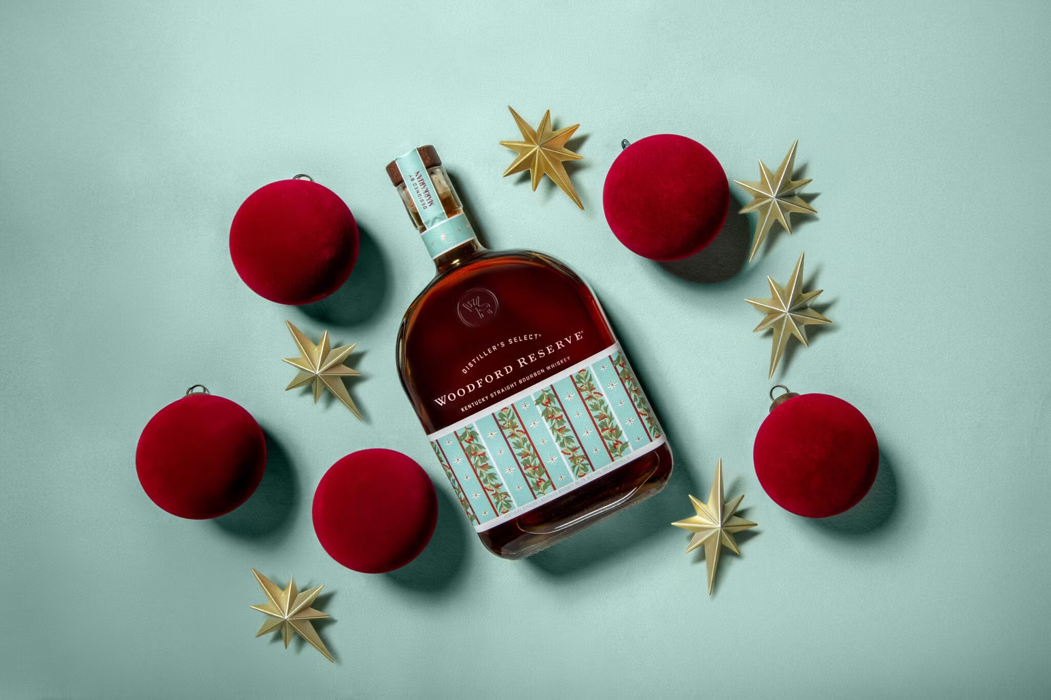 A bottle of Woodford Reserve Holiday Edition Bourbon 2025 “Designed by Markarian” is placed on a light blue background, surrounded by four red ornaments and six gold stars arranged neatly around it.